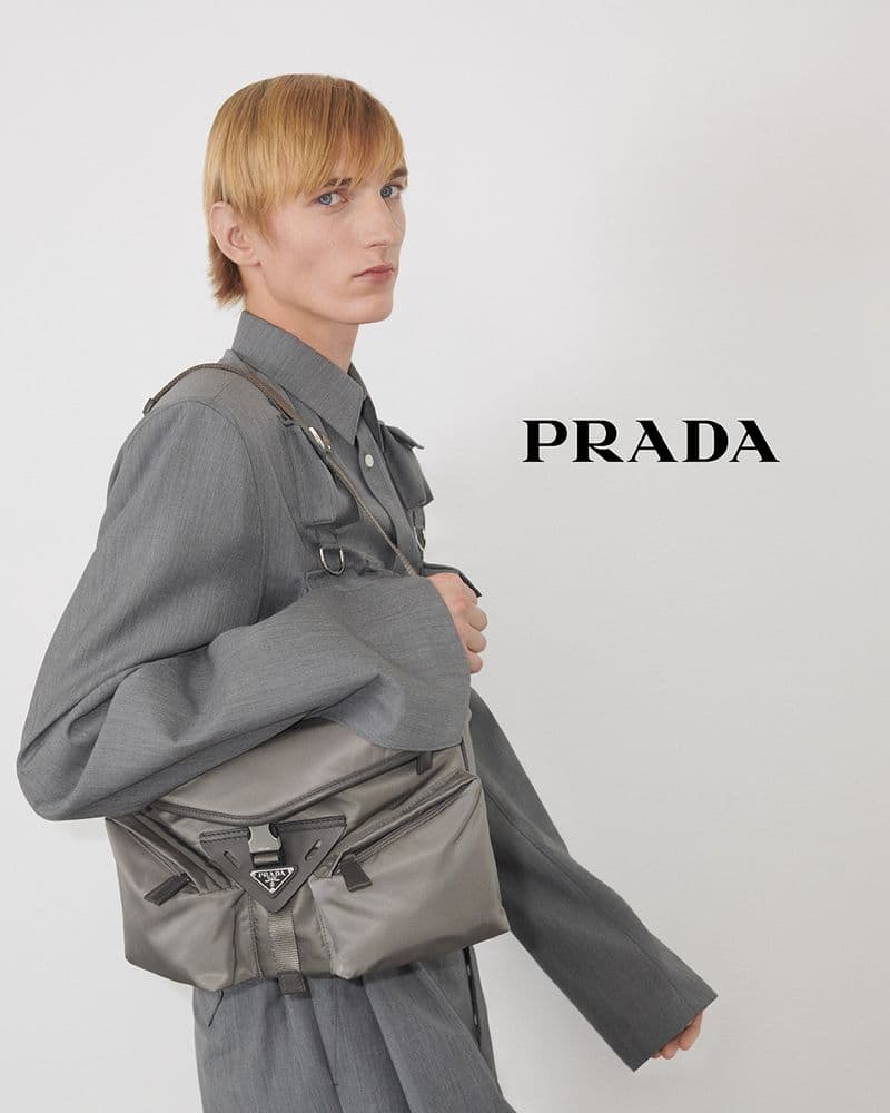 A light-skinned male model presents Prada's gray nylon bag, wearing matching gray jacket and shirt in a bright, minimalist setting.
