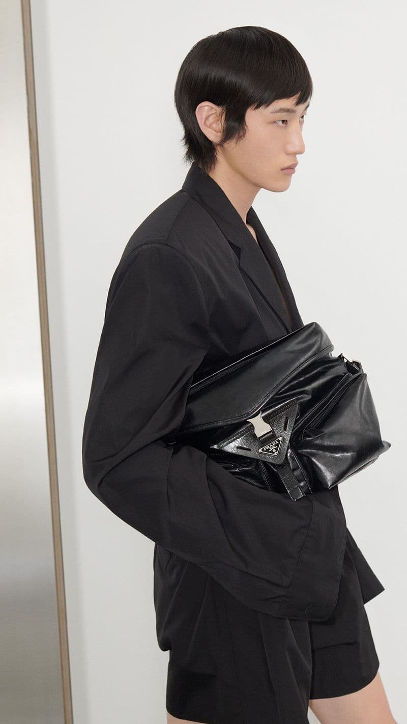 Model in black blazer holding a black leather bag against a plain white studio wall for Prada Spring 2024 ad campaign