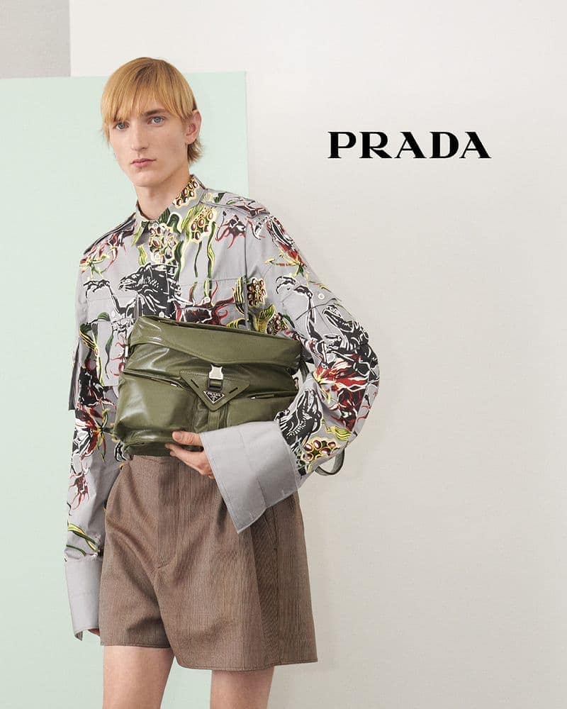 A blond male model poses against a white wall, holding a green Prada purse, wearing shorts, a printed shirt and gray background.