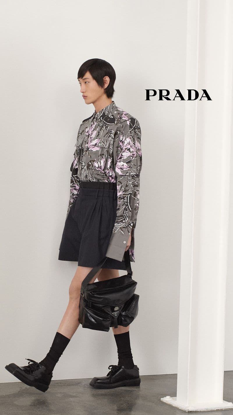 A person in a patterned shirt, black shorts, socks, and shoes poses with a black bag in front of a white wall with Prada logo.