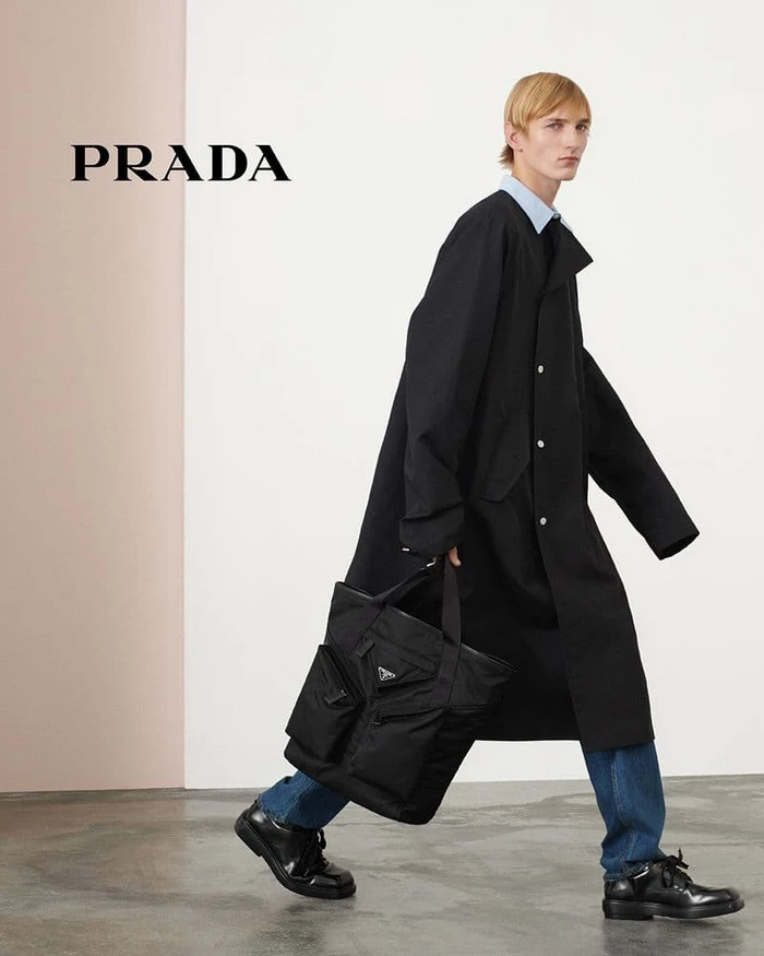 Model in black coat and blue jeans carrying a large black tote bag for Prada Spring 2024 campaign