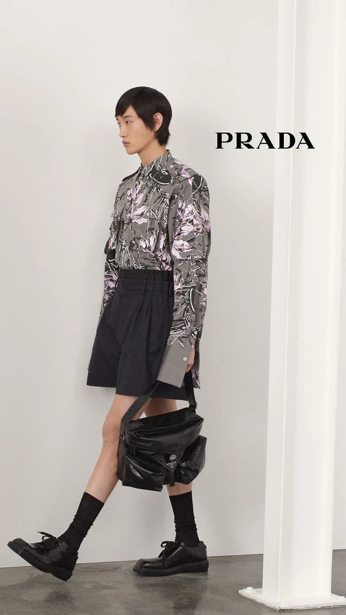 Model in printed long sleeve shirt and dark shorts walking past white column with Prada logo