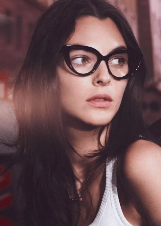 Portrait of a brunette woman wearing cat-eye glasses and a white tank top against a blurred background with some visible writing.
