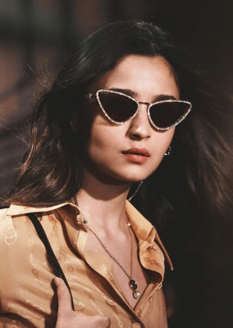 A woman poses in sunglasses and a yellow patterned blouse with a necklace. The photo is a close-up shot with a blurred background.
