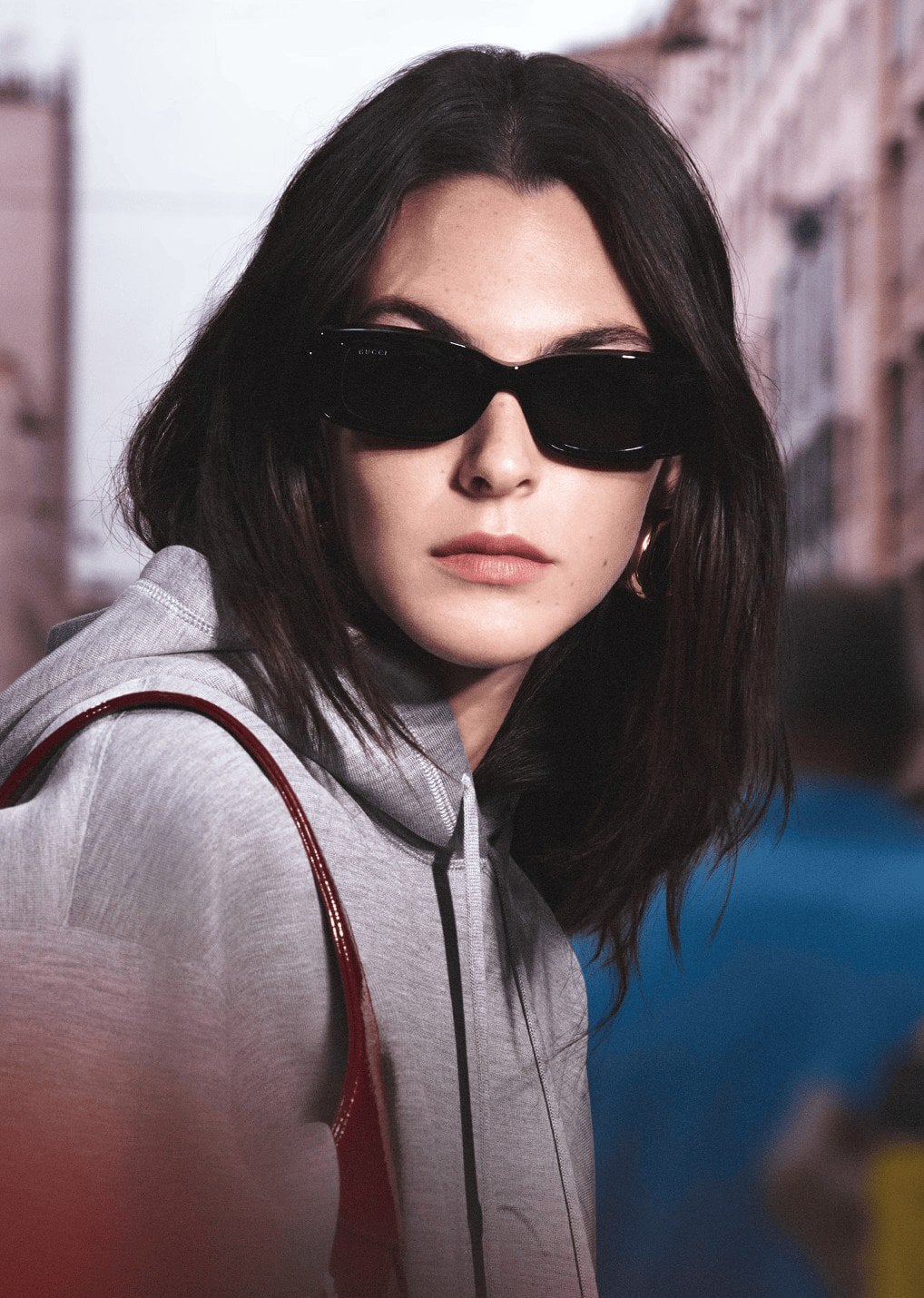 Brunette model in sunglasses and grey hoodie poses in an urban street scene. Fashion shot showcases modern casual wear and city lifestyle.