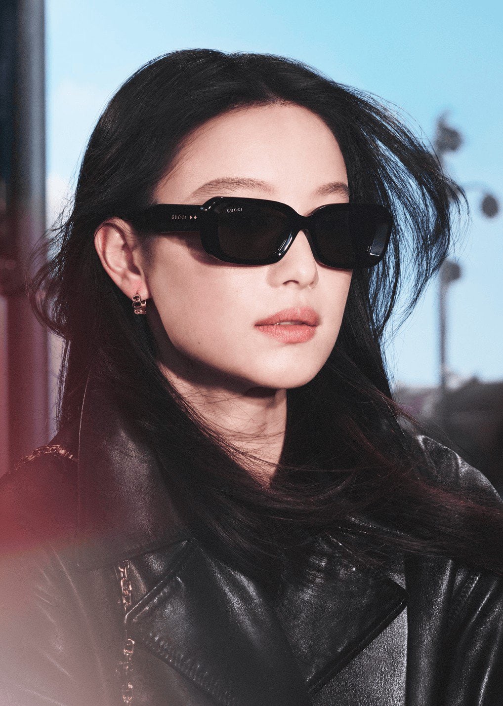 Woman with dark hair wearing black sunglasses and a leather jacket, standing outdoors with a blurred background of buildings.