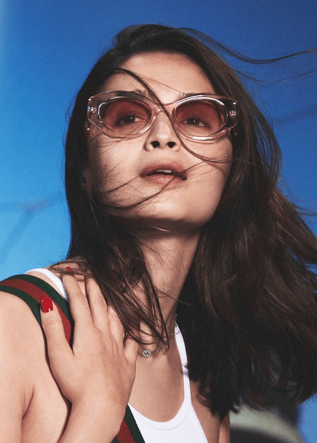 Close-up portrait of a stylish adult woman wearing pink sunglasses, a white top, and a necklace against a bright blue background.