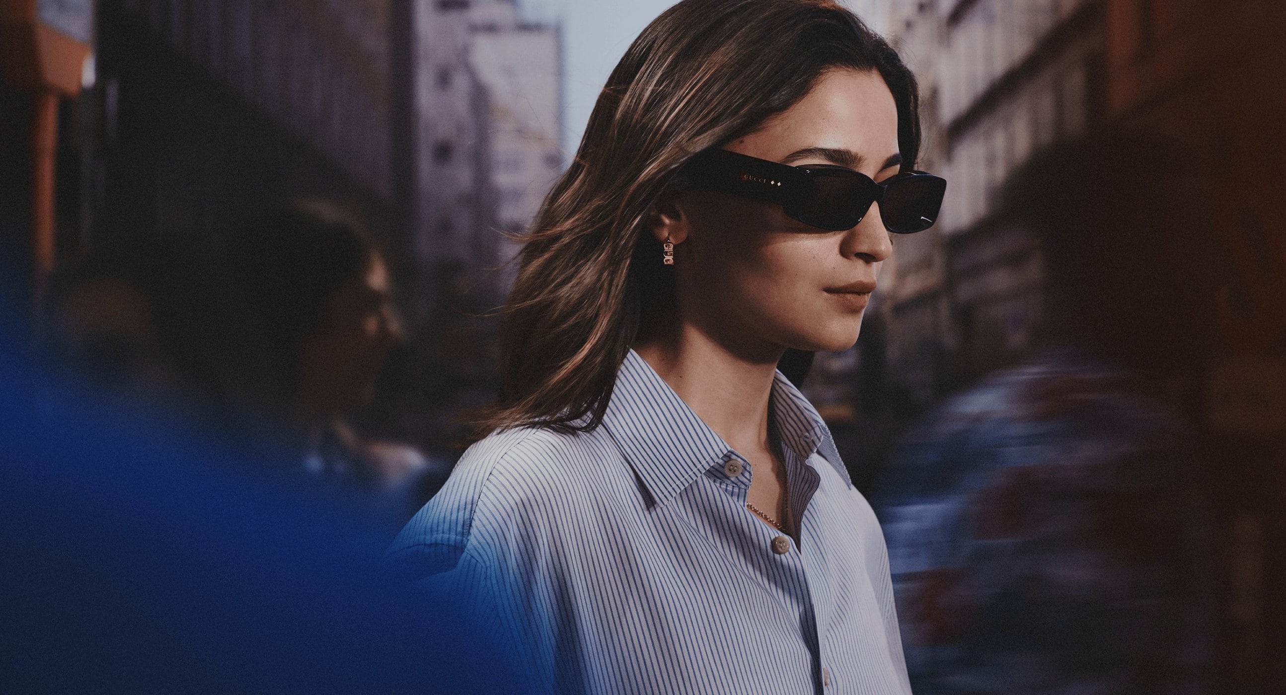 Brunette woman with sunglasses in striped shirt walks city street with blurred figures, buildings in background, fashion editorial.
