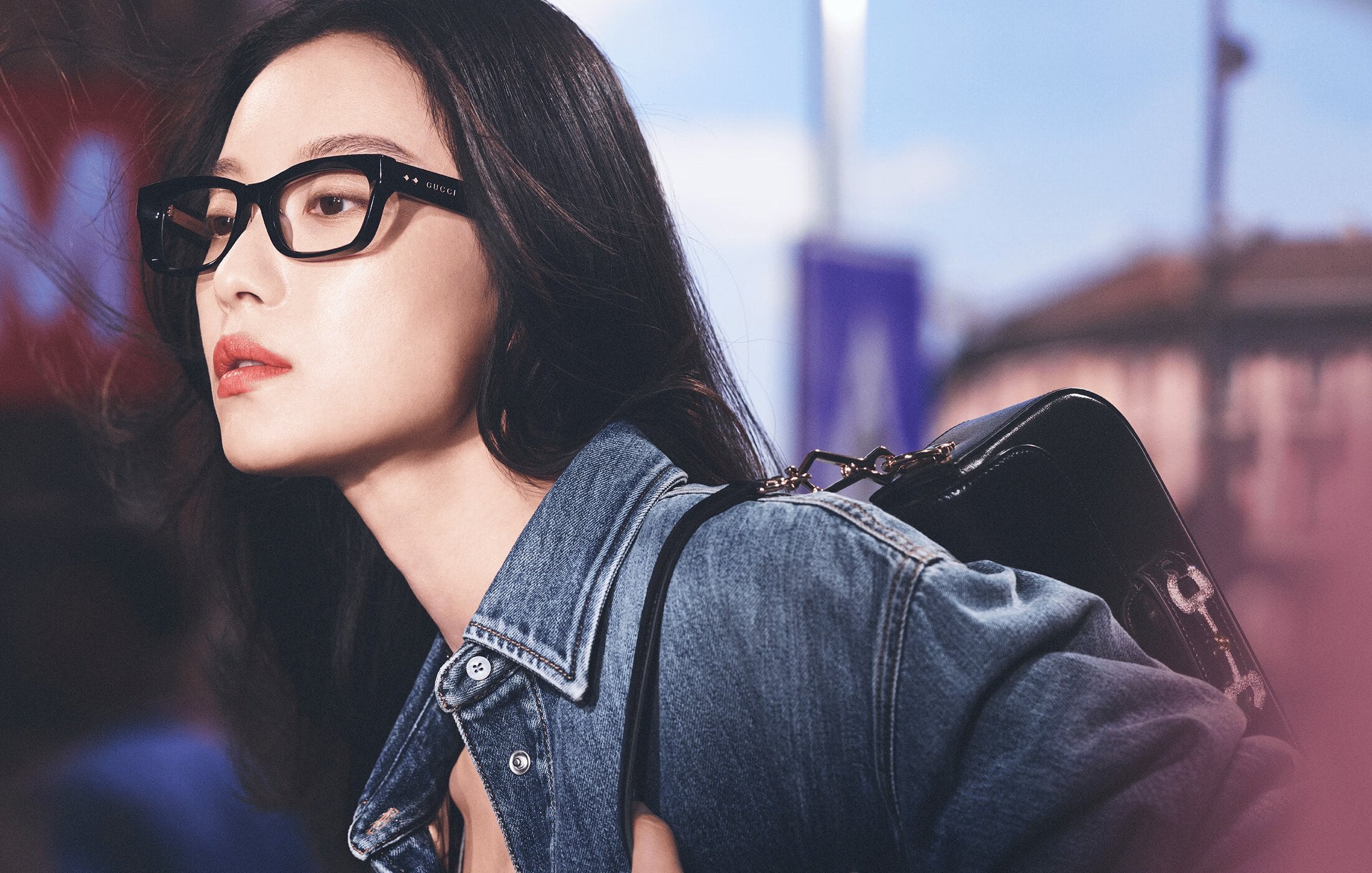 An Asian woman with long dark hair poses in a denim jacket and black-framed glasses, accessorized with a black purse in an outdoor setting.