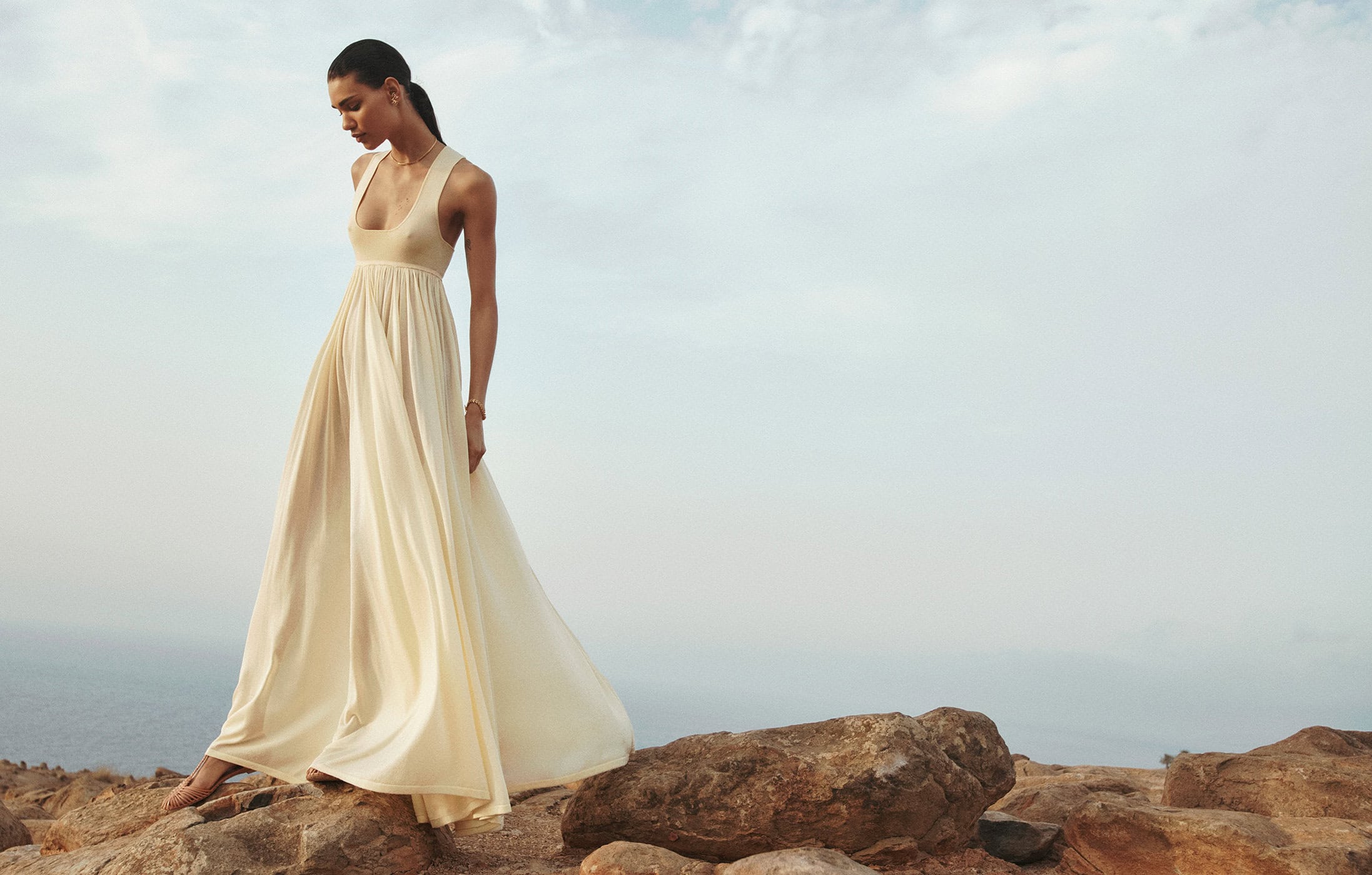 A woman in a flowing white dress stands on rocks overlooking the sea under a bright cloudy sky in a fashion or lifestyle image.