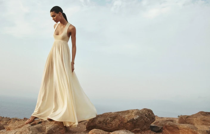 A woman in a flowing white dress stands on rocks overlooking the sea under a bright cloudy sky in a fashion or lifestyle image.
