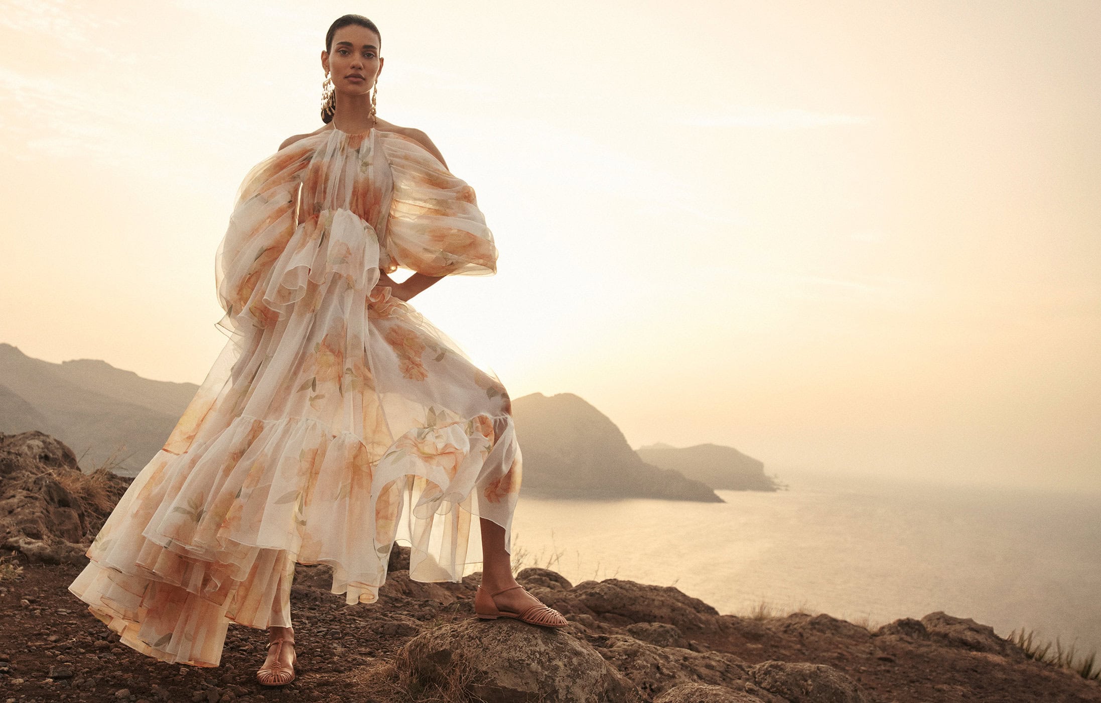 Elegant adult female model in a flowing chiffon gown with a floral pattern poses outdoors on a rocky terrain with a coastal background.