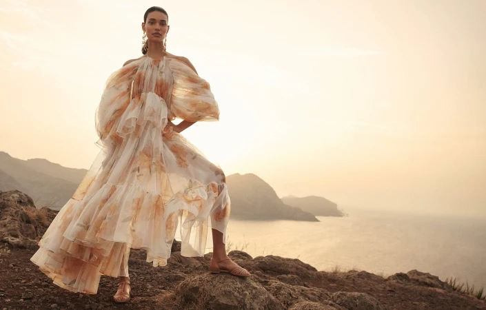 Elegant adult female model in a flowing chiffon gown with a floral pattern poses outdoors on a rocky terrain with a coastal background.