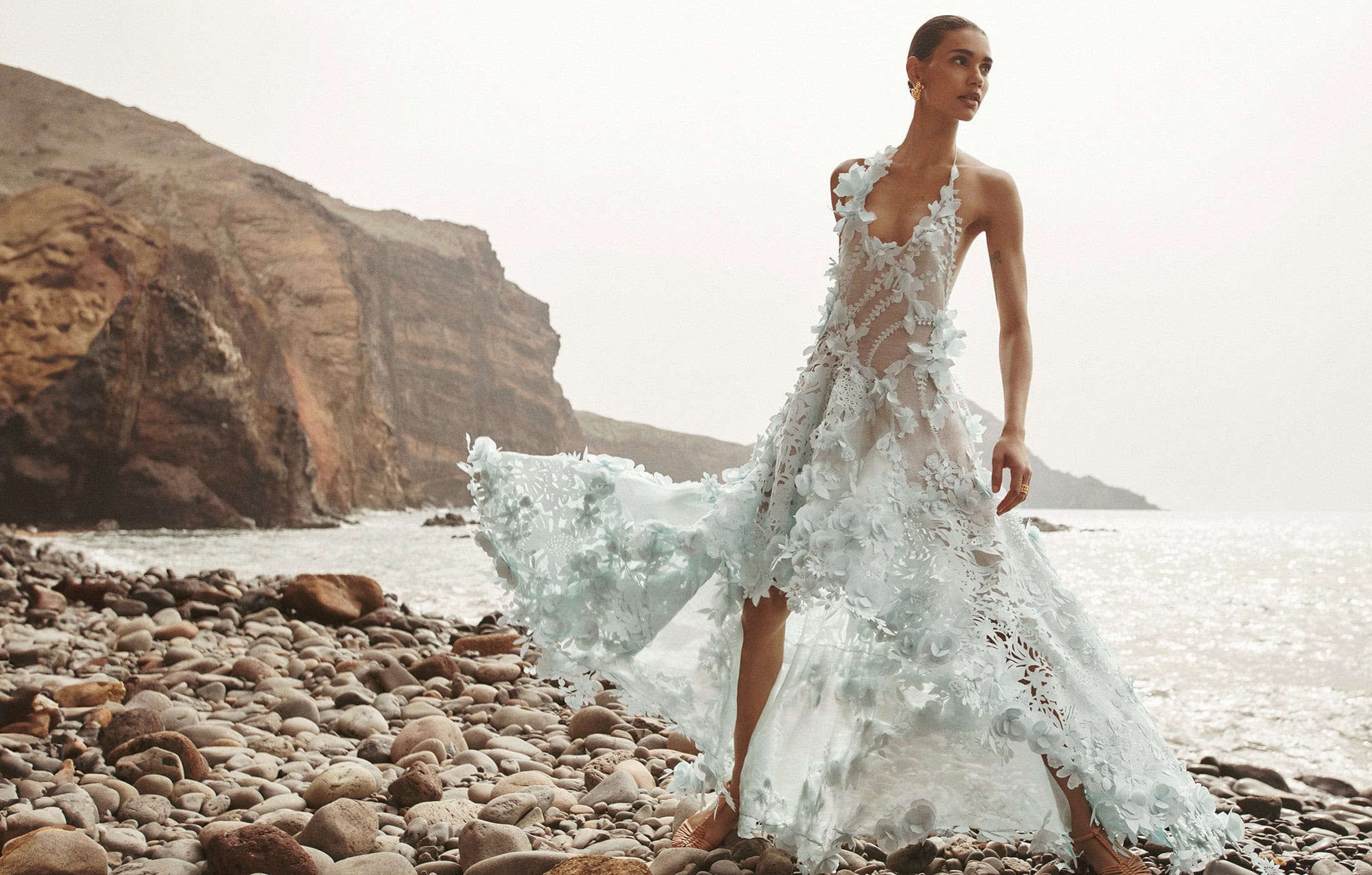 Woman in couture gown with floral accents poses on a rocky beach with cliffs and ocean for fashion editorial photography.