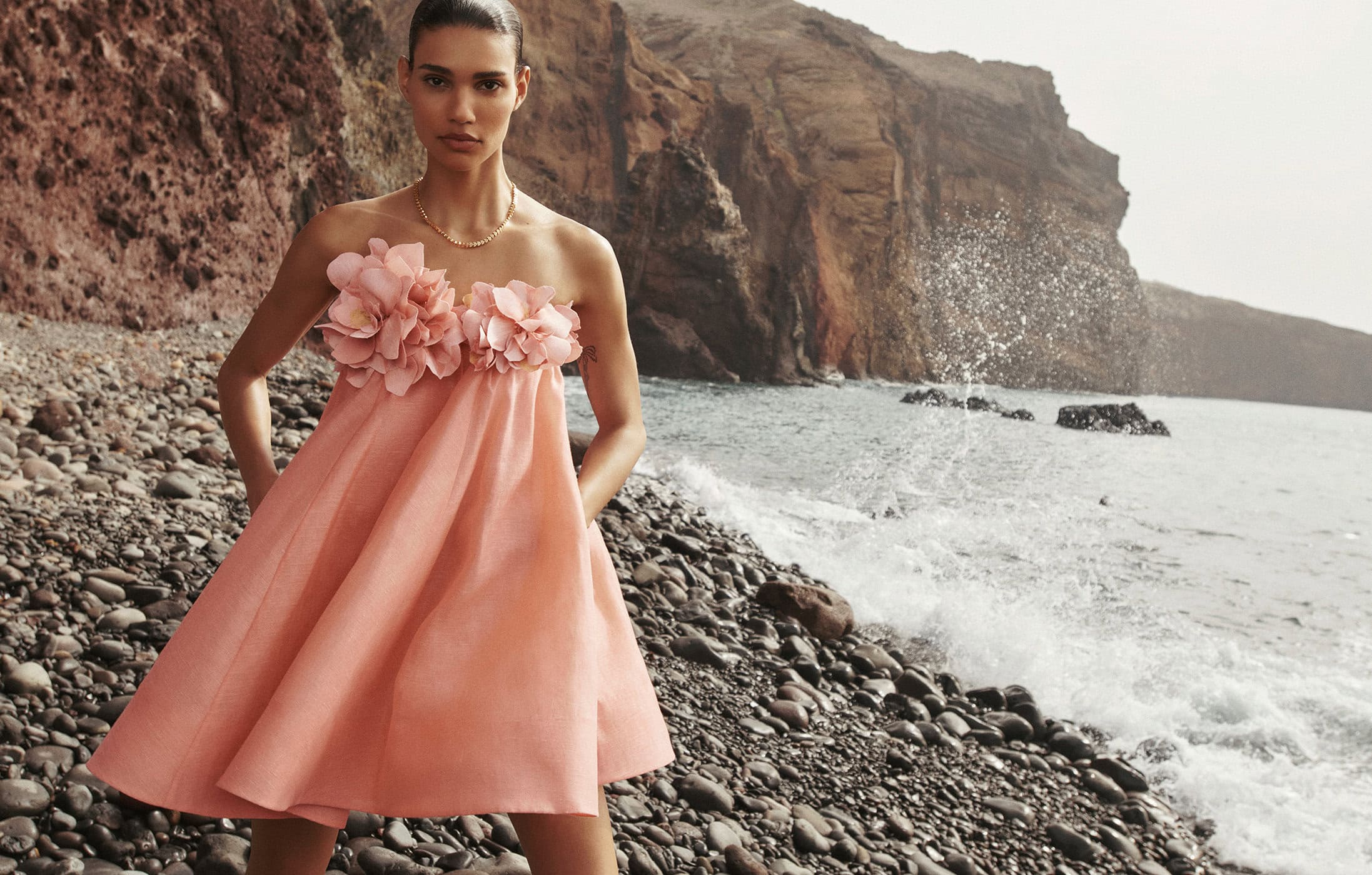 Woman in pink dress with floral detail poses on a rocky beach with ocean and cliffside in the background, fashion editorial outdoor shot.
