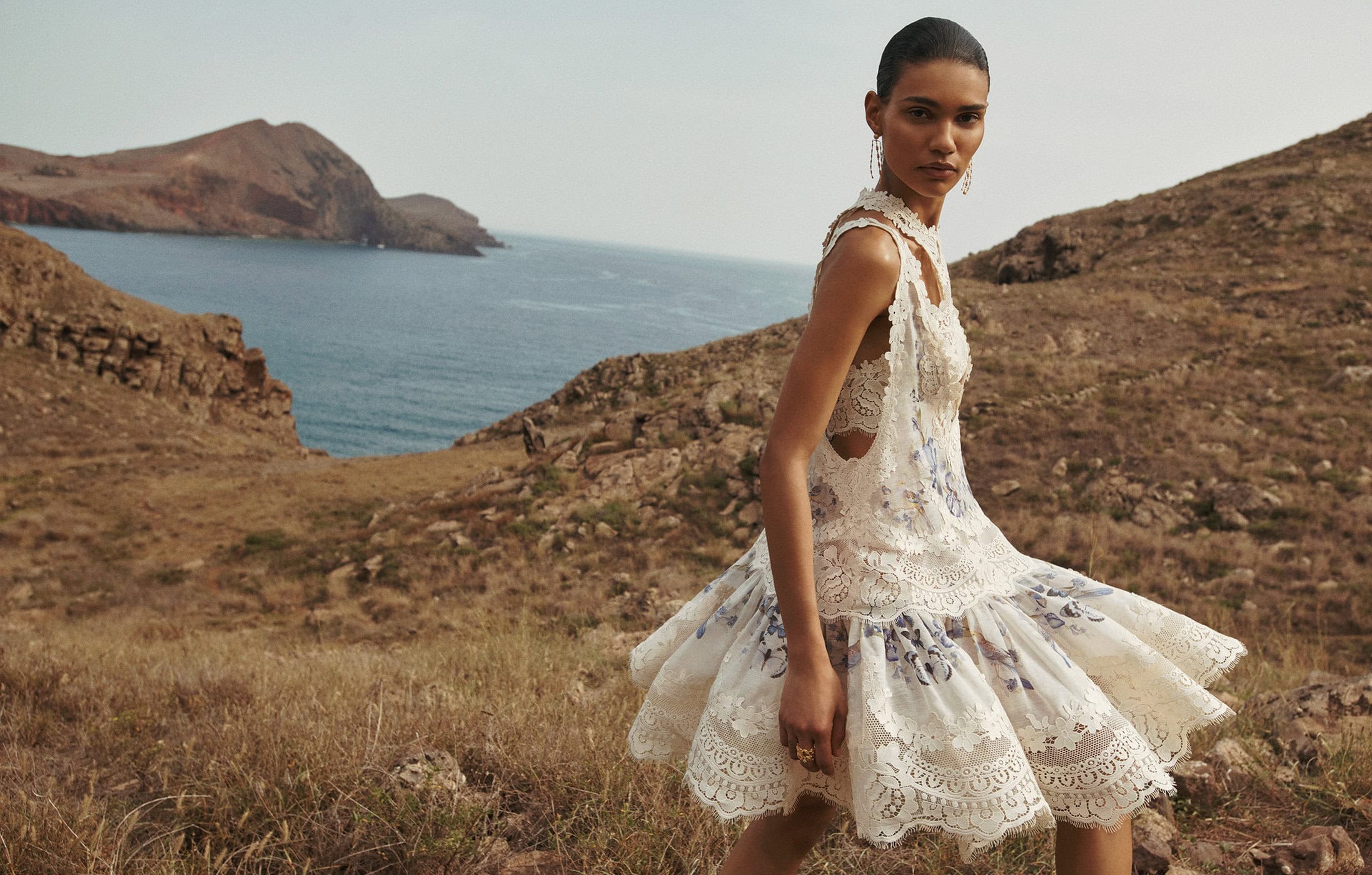An adult female model is wearing a lacy, floral dress in an outdoor fashion editorial shoot with rocky cliffs and blue ocean in the background.