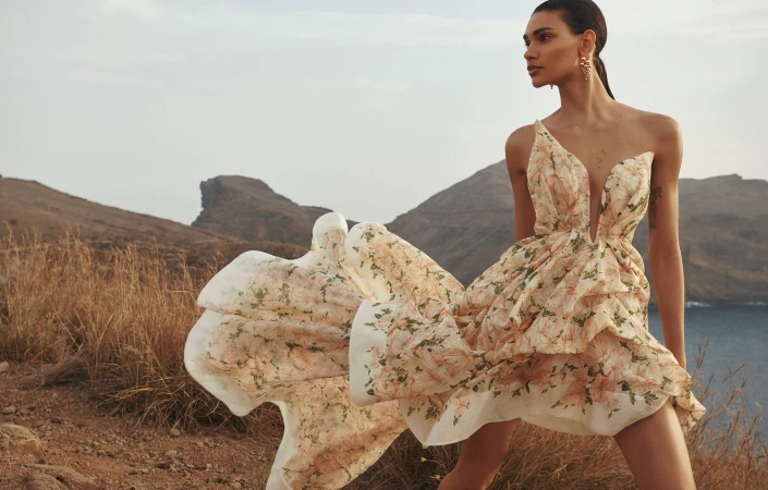A young adult woman poses in a flowing floral dress with a dramatic mountain backdrop. Editorial fashion photography in natural light.