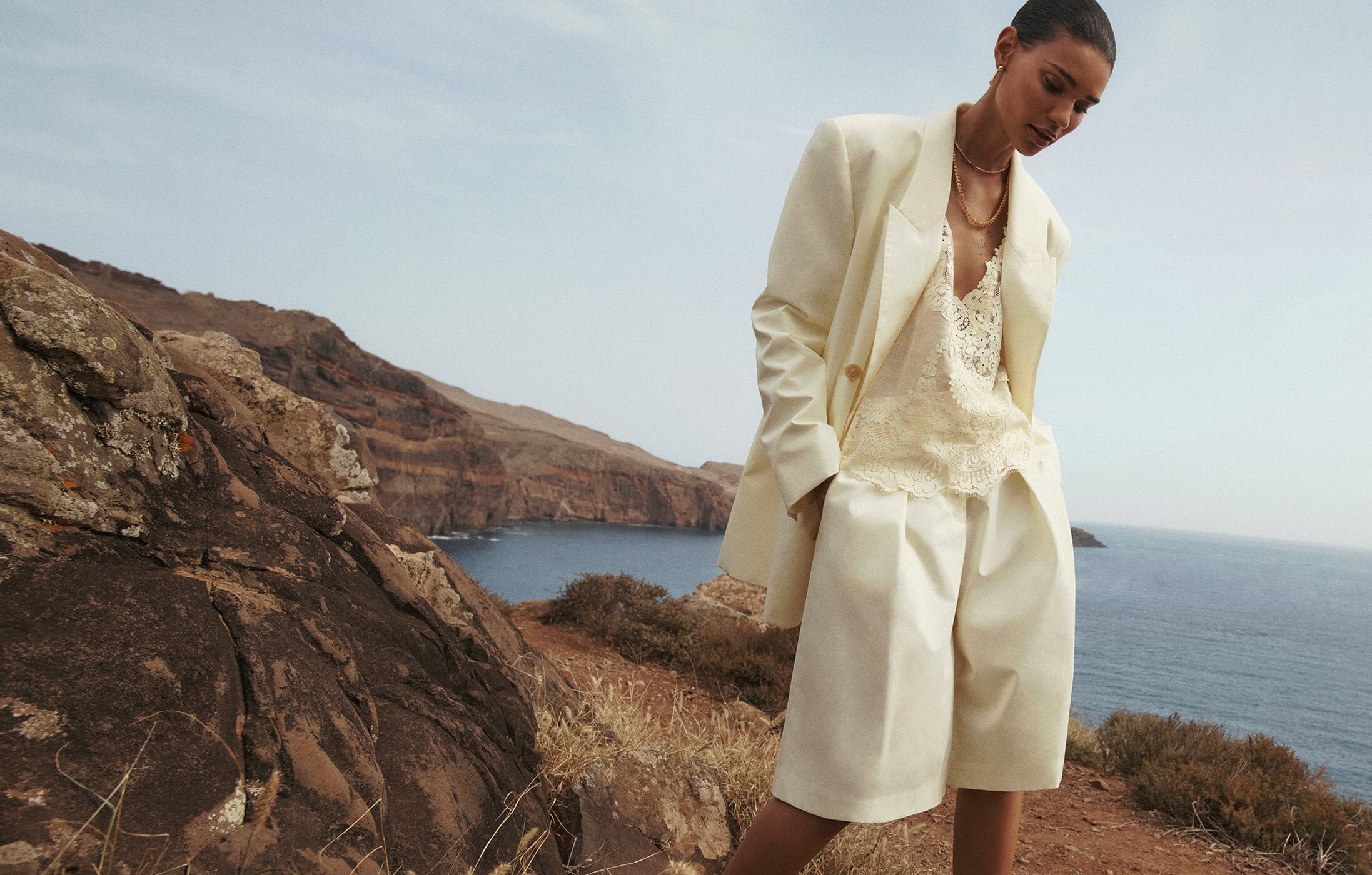 Woman in cream blazer and shorts posing on rocky coast with ocean view. Fashion shot with natural light and scenic background.