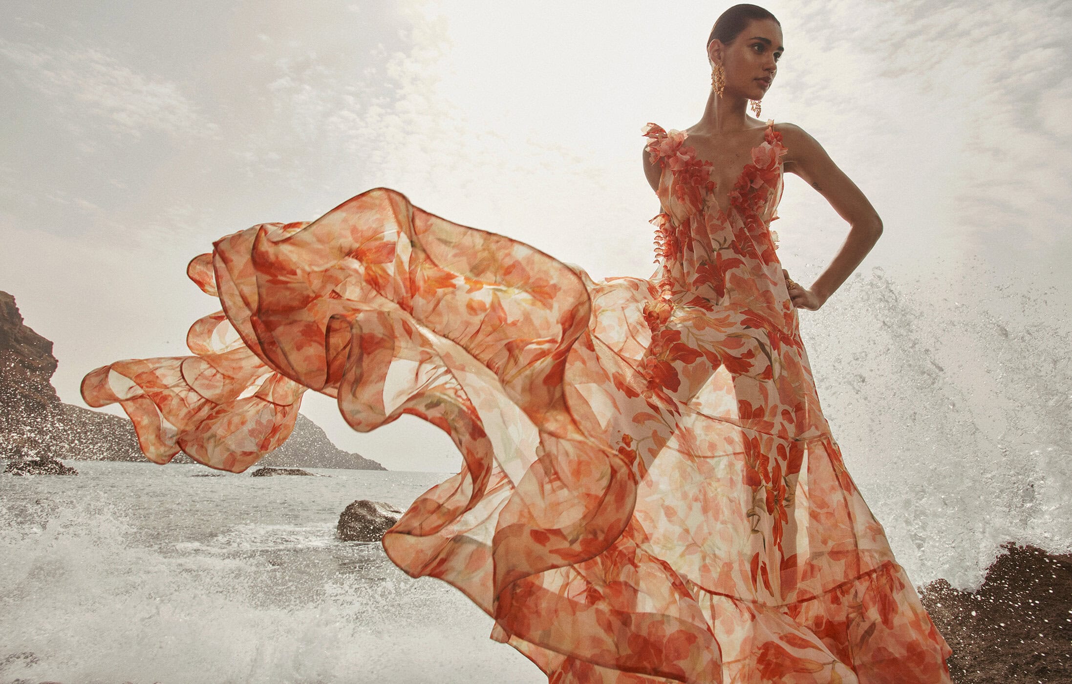 Woman in a flowing floral dress stands by the ocean as waves crash around the rocks in a fashion-forward outdoor shot.