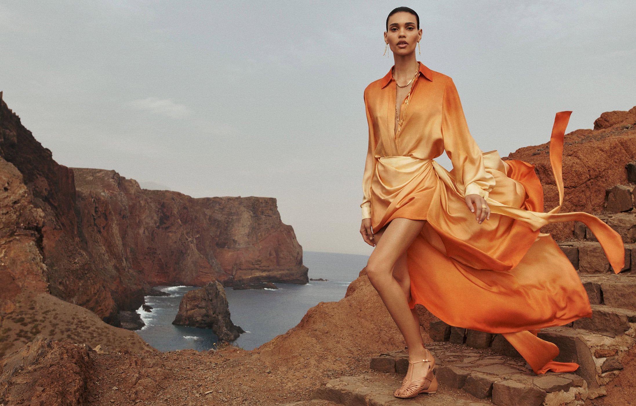 An adult female fashion model is walking on stone stairs on a rocky cliffside overlooking the ocean, wearing an orange dress.