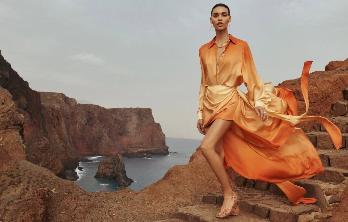 An adult female fashion model is walking on stone stairs on a rocky cliffside overlooking the ocean, wearing an orange dress.