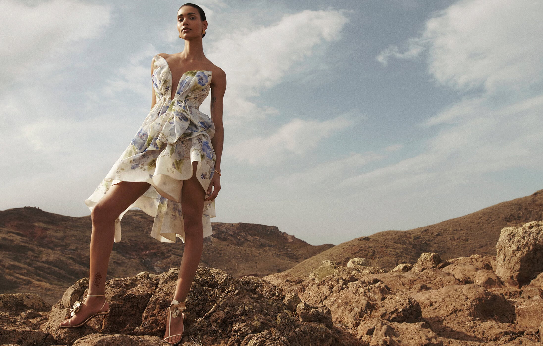 Woman in floral dress stands on rocks against a partly cloudy sky and distant mountains, wearing ankle-strap heeled sandals outdoors.