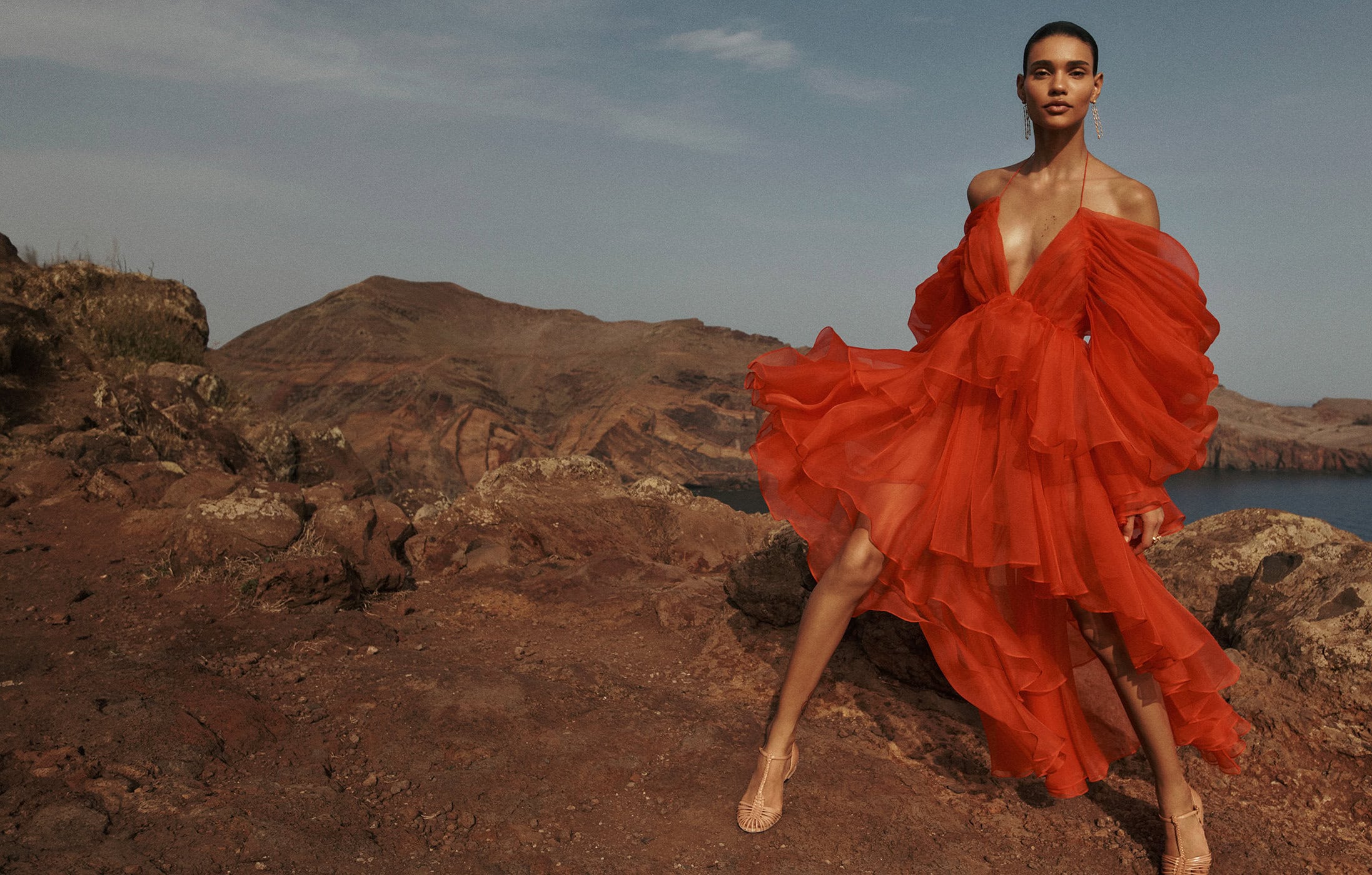 Fashion shot of a young adult woman wearing a red dress, standing on rocky terrain with mountains and the ocean in the background.