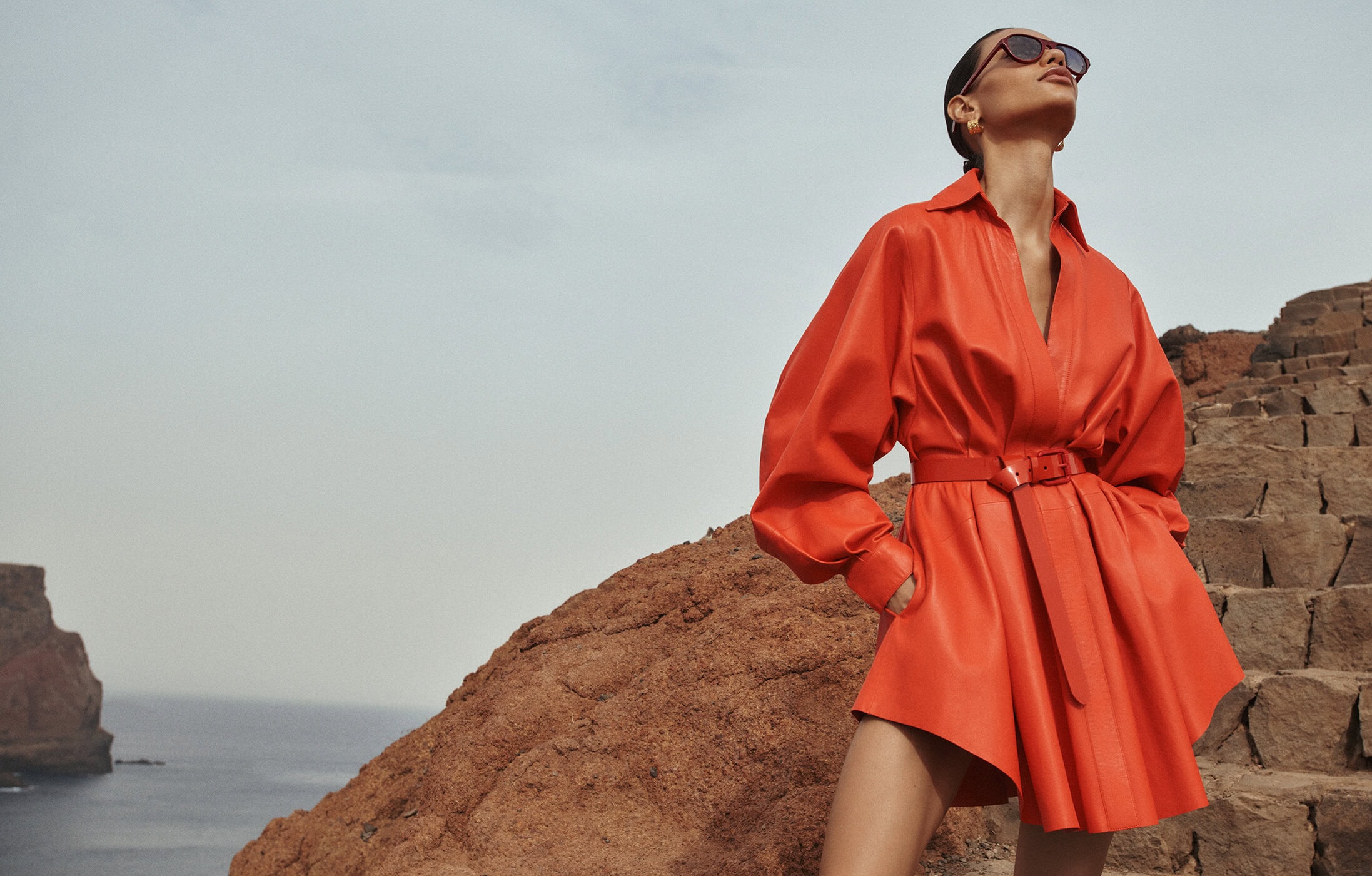 A woman in a red dress and sunglasses stands on a rocky outcrop overlooking the ocean, posing against a cloudy sky.