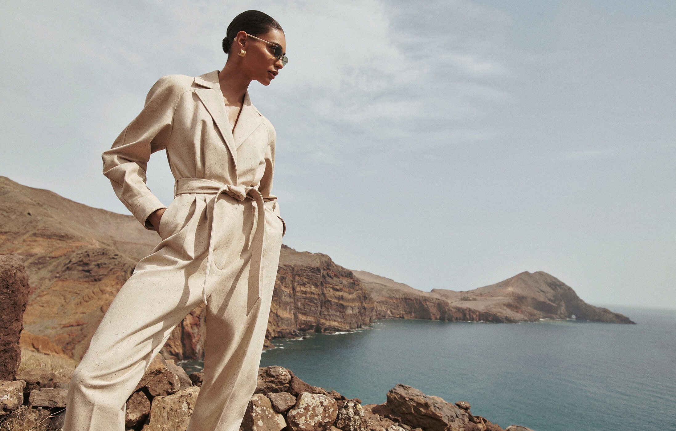Woman in a beige jumpsuit with sunglasses poses on a rocky outcrop overlooking the ocean and landscape on a sunny day.