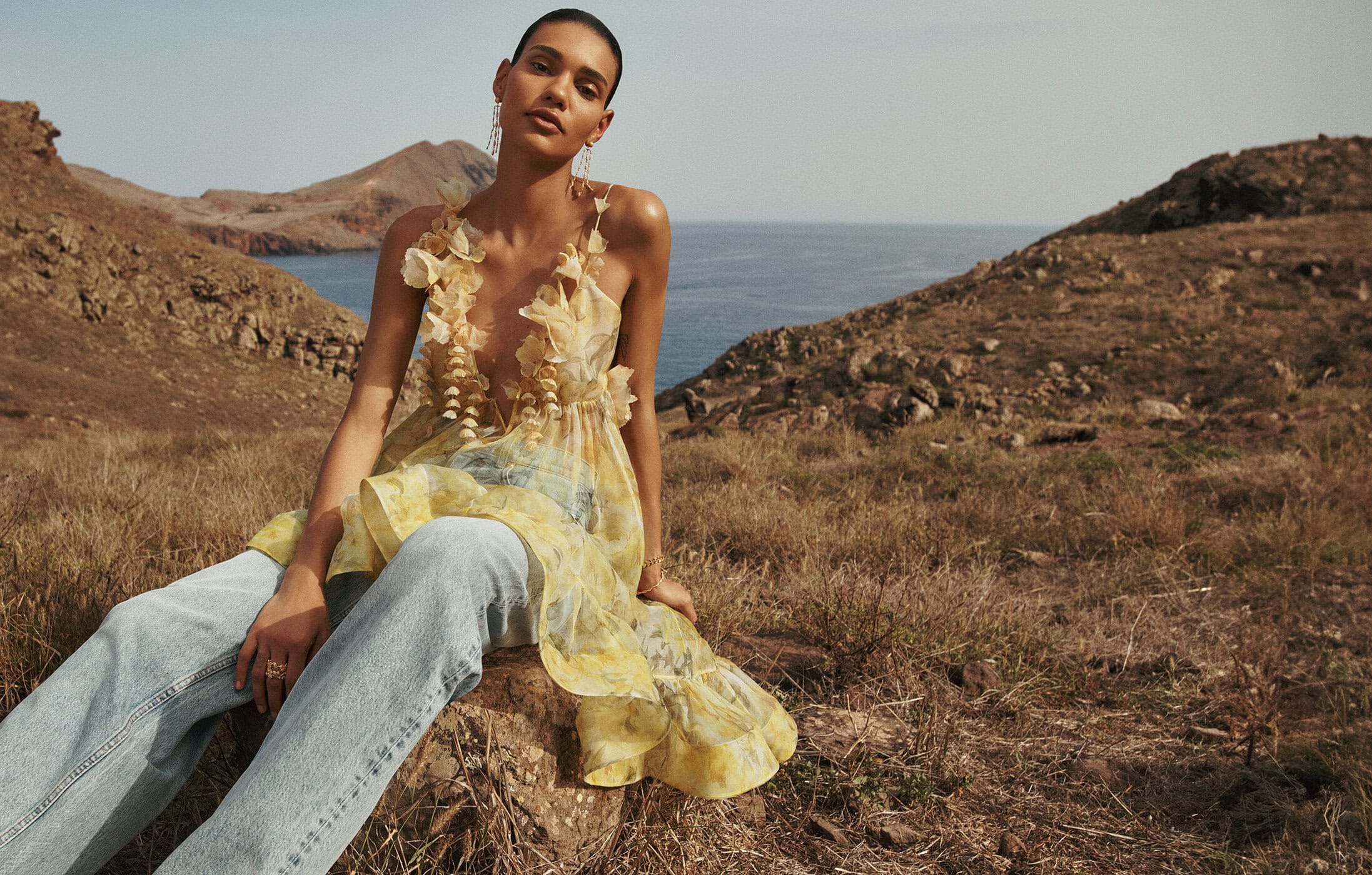Fashionable adult woman sits in a ruffled top and jeans against an outdoor landscape with hills and the ocean in the background.