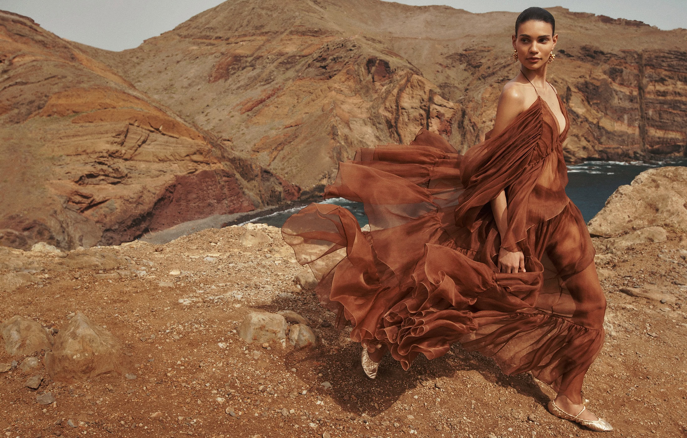 Woman in flowing brown dress on a rocky cliff with mountains and ocean in the background, fashion editorial outdoor shot.