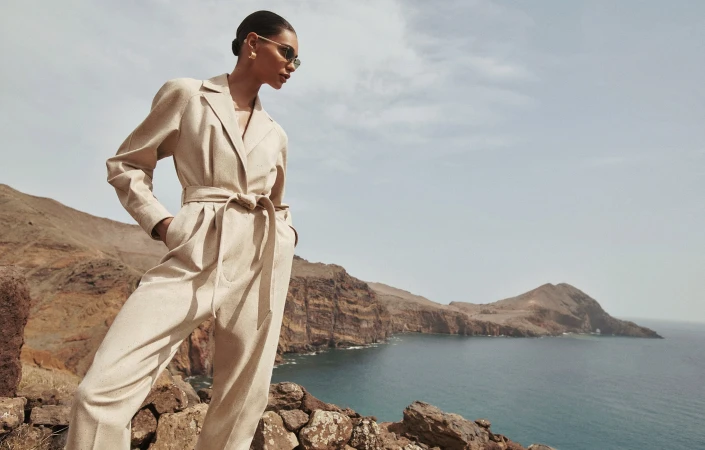 Model in beige belted jumpsuit standing on rocky coastal cliffs above ocean for Zimmermann campaign