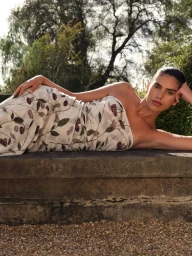 Model Sun Mizrahi lying on a low stone wall wearing a strapless patterned dress against trees