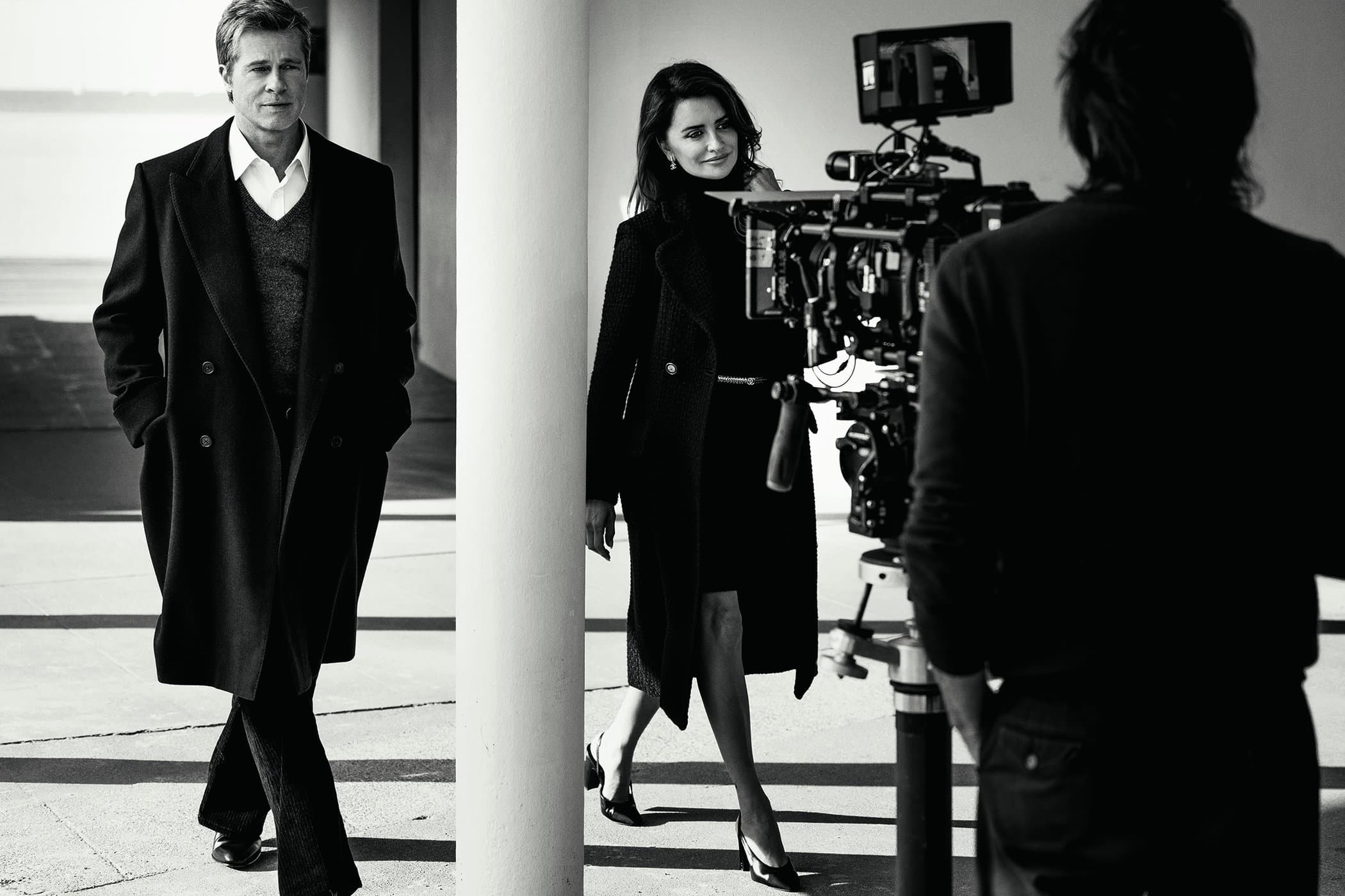 Black and white fashion shot featuring Brad Pitt and Penelope Cruz with a film crew, showcasing elegant overcoats in an outdoor setting.