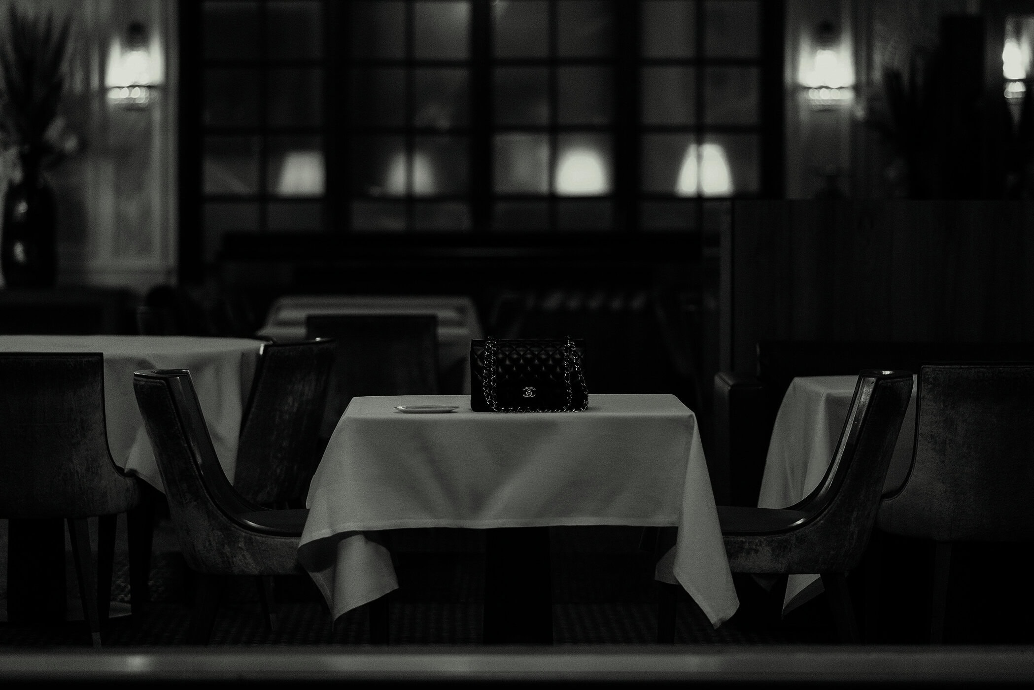 Chic black Chanel handbag on a linen-covered table in a softly lit interior, complemented by dark wood chairs and vintage windows.