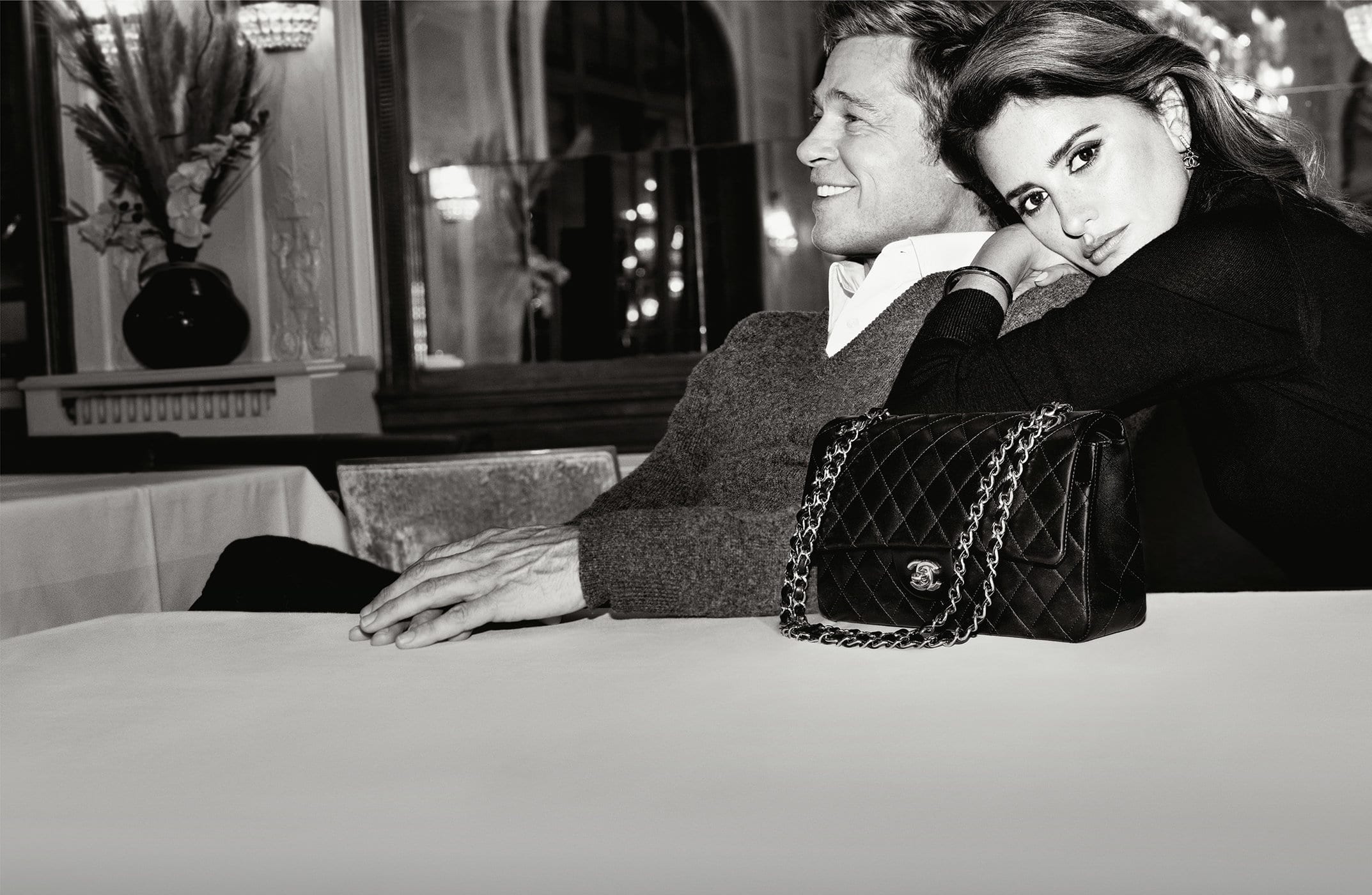 Brad Pitt and Penelope Cruz pose next to a Chanel quilted handbag in a dimly lit indoor setting, showcasing a blend of elegance and sophistication.