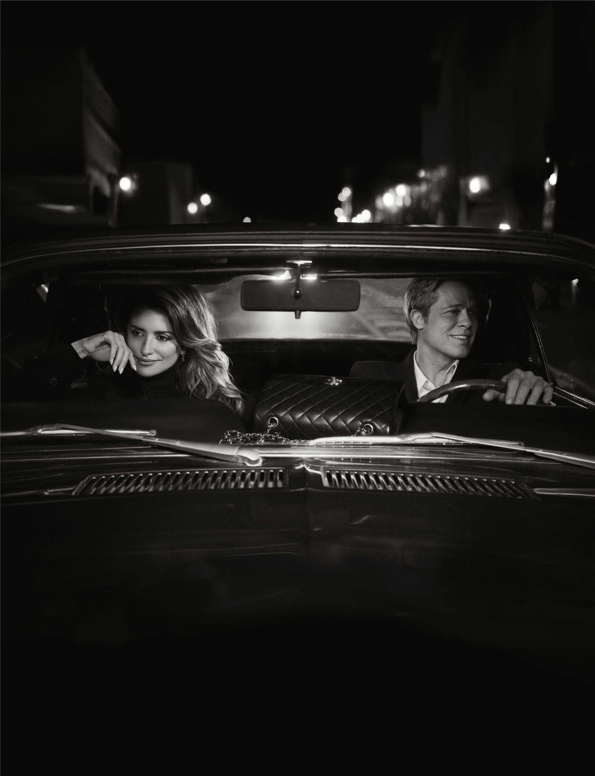 Monochrome night shot of a smiling couple in a vintage car with a quilted handbag on the dashboard, city lights in background.