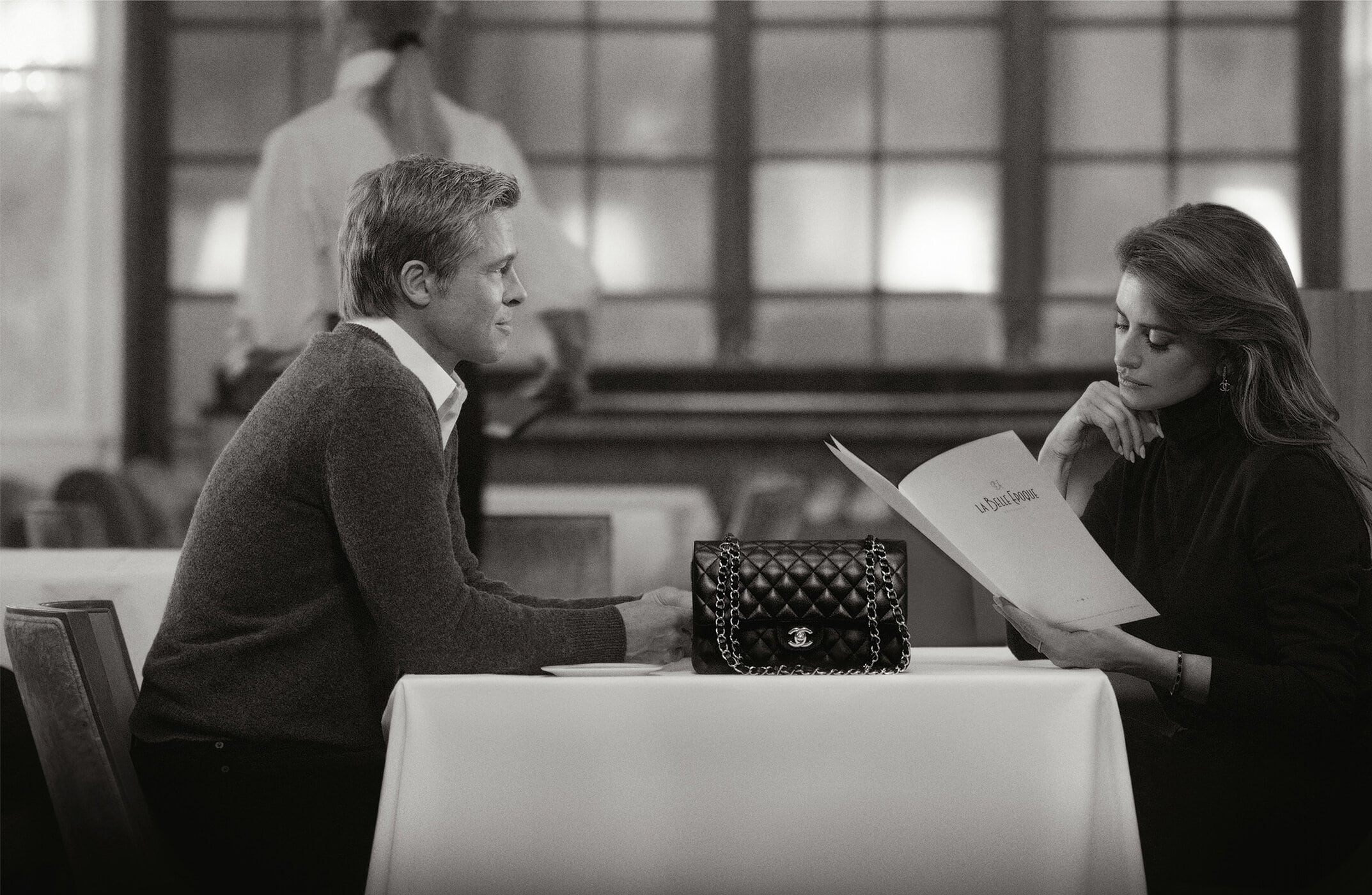 Black and white image of Brad Pitt and Penelope Cruz at a table with a Chanel handbag in a restaurant, reading a menu.