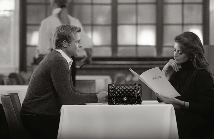 Black and white image of Brad Pitt and Penelope Cruz at a table with a Chanel handbag in a restaurant, reading a menu.