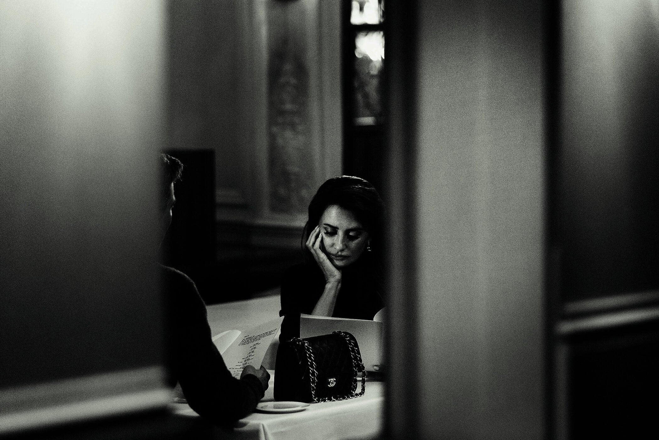 Penelope Cruz seated at a table with Brad Pitt in a monochromatic interior for the Chanel Iconic Handbag campaign.