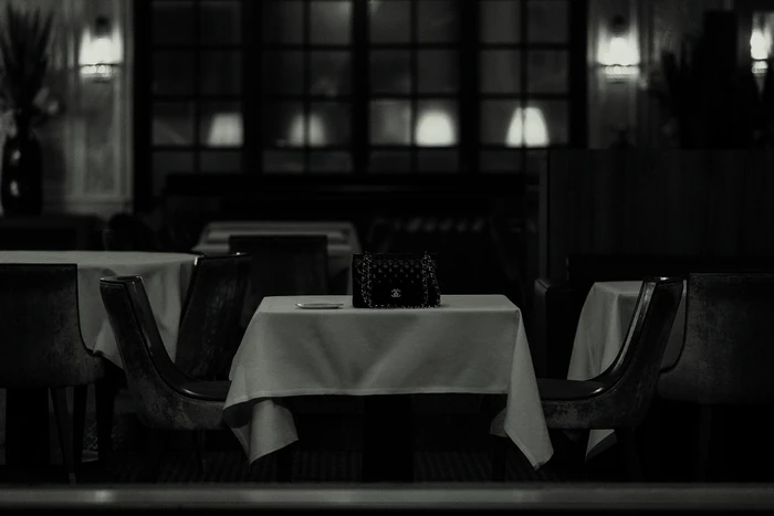Black and white image of a Chanel handbag resting on a white tablecloth-covered table inside an interior restaurant setting.