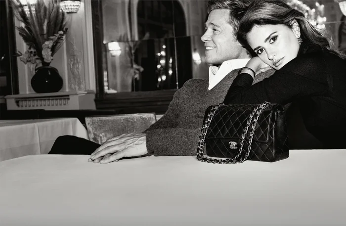 Brad Pitt and Penelope Cruz seated together at a table in black and white with a classic Chanel quilted handbag in the foreground.