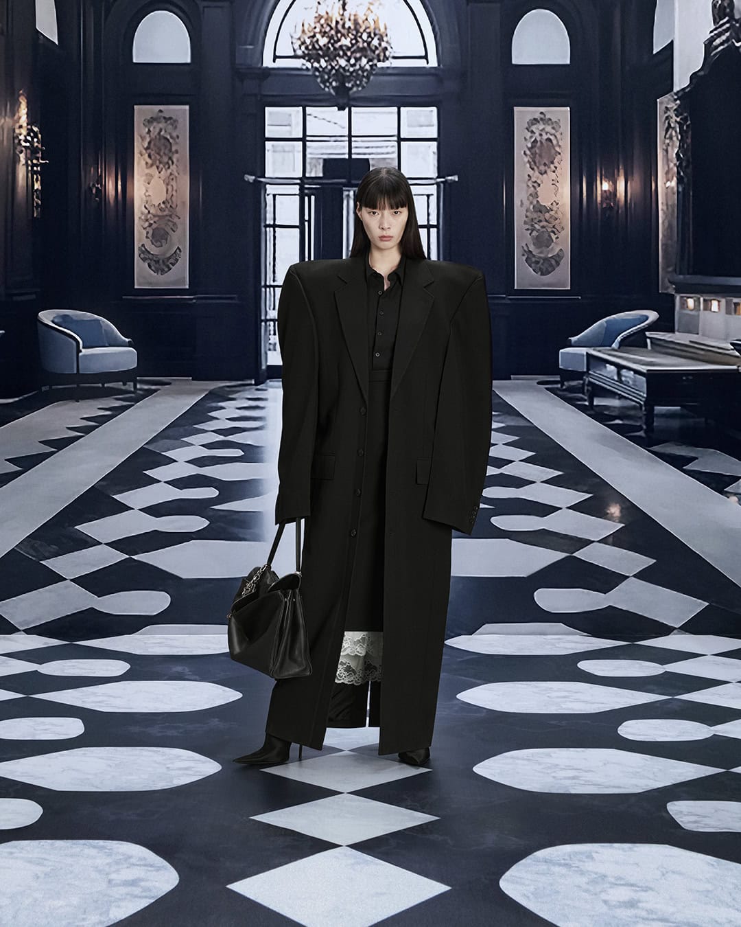 A stylish adult female poses in a black oversized coat with a handbag, set against a chic black and white checkered floor indoors.