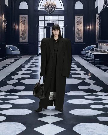 A stylish adult female poses in a black oversized coat with a handbag, set against a chic black and white checkered floor indoors.