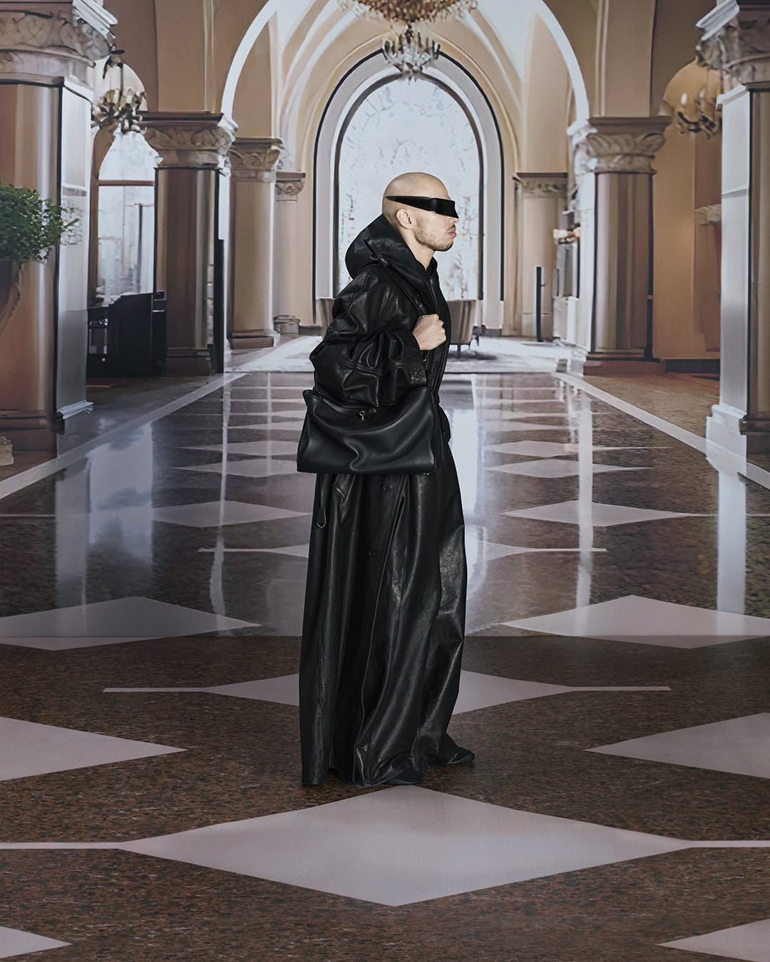 A bald adult male, wearing black visor sunglasses and a draped black leather outfit with a matching bag, stands in an arched hallway.