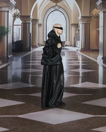 A bald adult male, wearing black visor sunglasses and a draped black leather outfit with a matching bag, stands in an arched hallway.