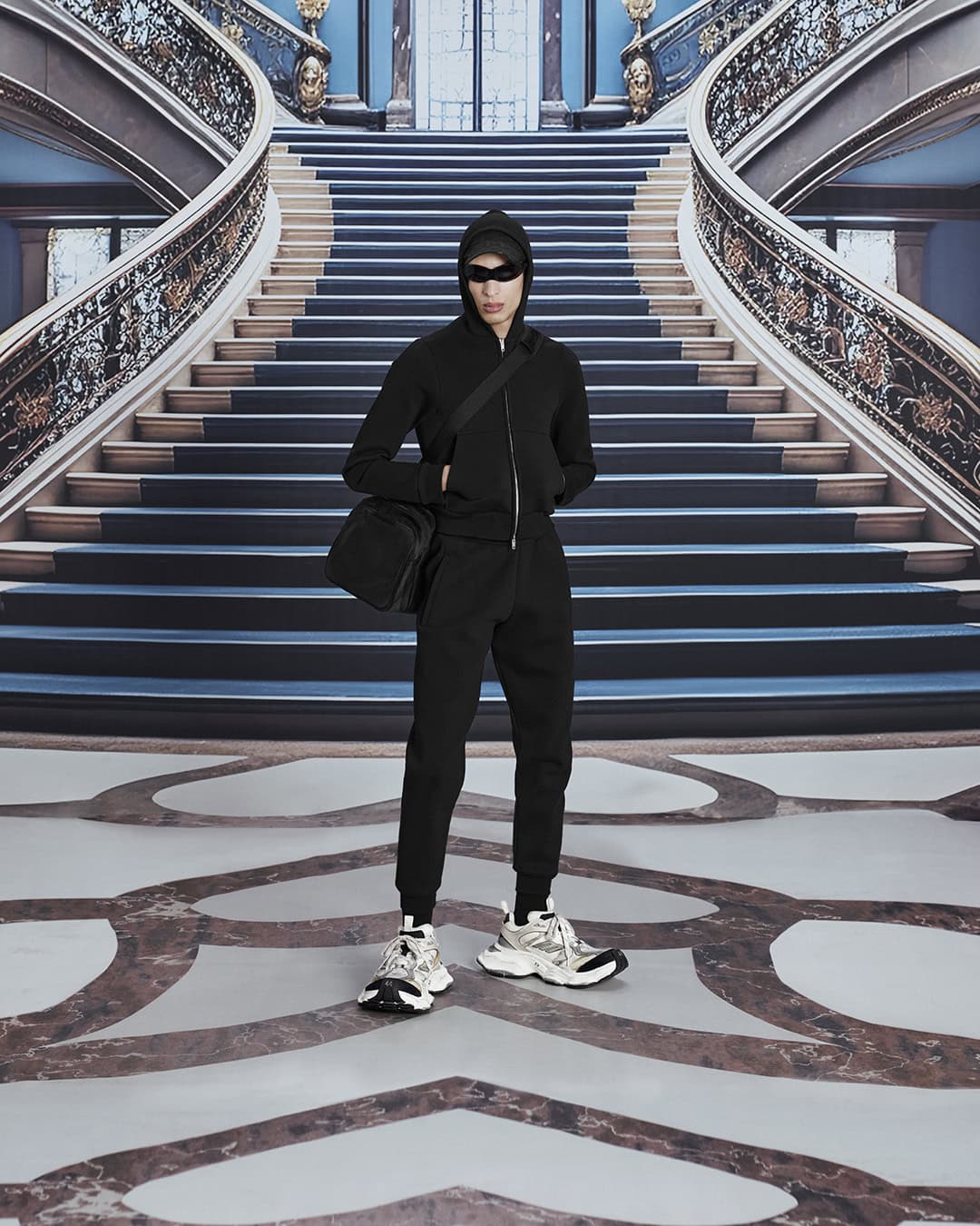 Person in black hooded tracksuit with shoulder bag and sneakers stands against a grand staircase backdrop in a fashion editorial.
