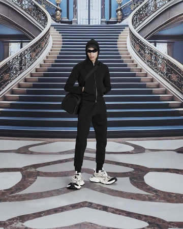 Person in black hooded tracksuit with shoulder bag and sneakers stands against a grand staircase backdrop in a fashion editorial.
