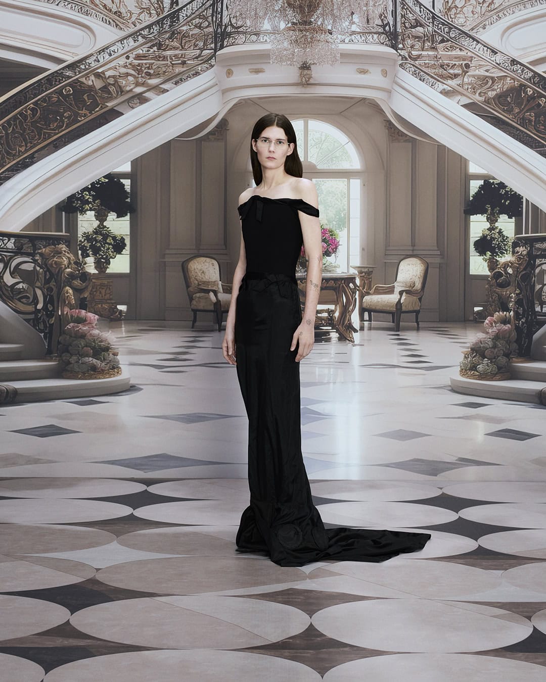 Elegant woman in off-the-shoulder gown poses in a grand marble hall with a curving staircase and ornate chandelier, fashion editorial.