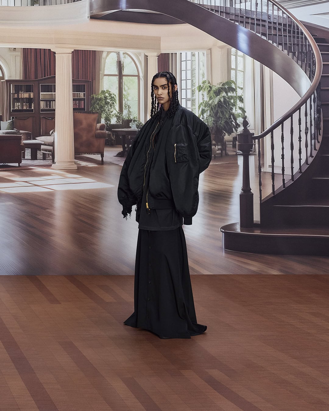 Adult female poses in an oversized black bomber jacket and black skirt inside a building with a staircase and decorative background.